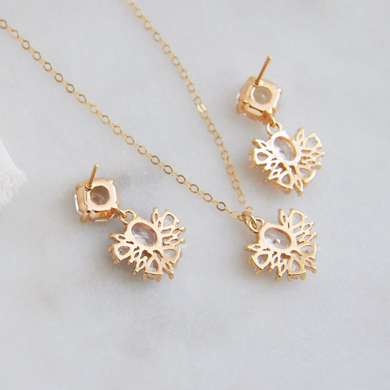 May include: A gold-colored jewelry set featuring a necklace and a pair of earrings. Each piece has a square-shaped top with a dangling floral design, all adorned with clear gemstones. The set is displayed on a white surface.