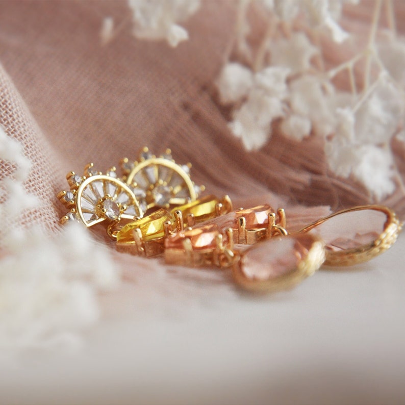 May include: Close-up of several pieces of jewelry with gold settings and colorful gemstones. The gemstones are in shades of yellow, orange, and pink. Some pieces have a half-circle design with clear stones. The jewelry is arranged on a soft, pink fabric.