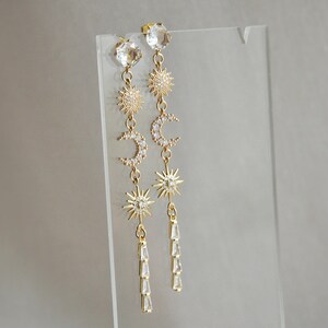 May include: A pair of gold-colored celestial-themed earrings. Each earring features a large clear crystal at the top, followed by a sunburst, a crescent moon, another sunburst, and a dangling cluster of clear crystals. The earrings are displayed on a clear stand.