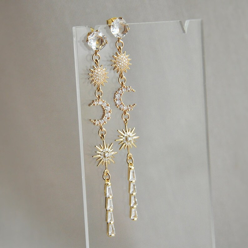 May include: A pair of gold-colored celestial-themed earrings. Each earring features a large clear crystal at the top, followed by a sunburst, a crescent moon, another sunburst, and a dangling cluster of clear crystals. The earrings are displayed on a clear stand.