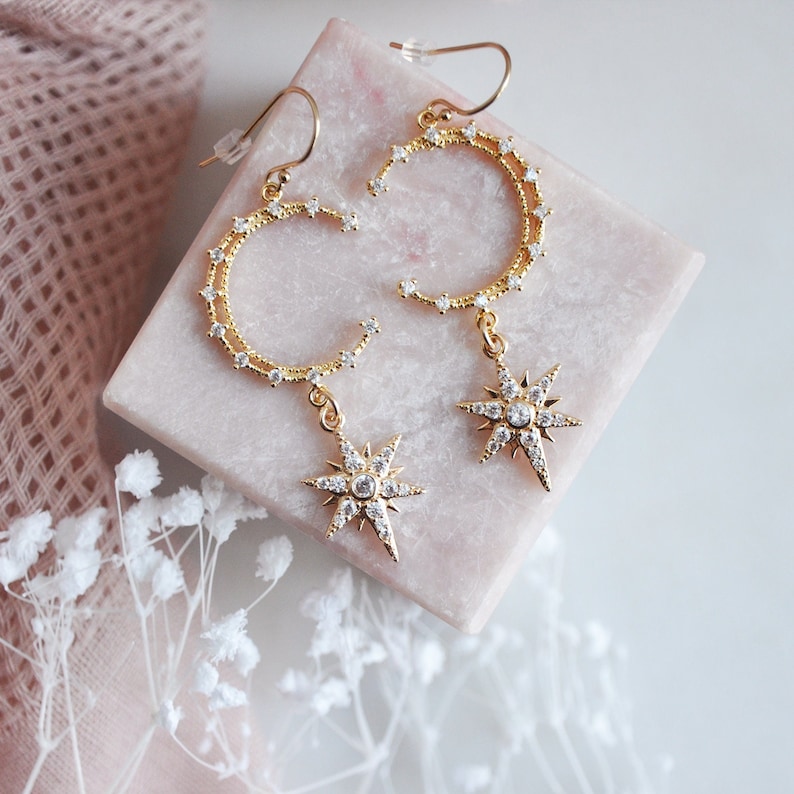 May include: Gold-toned celestial earrings featuring crescent moon and star pendants. The earrings are adorned with small, sparkling crystals. The earrings are displayed on a pink square surface, with a soft pink fabric in the background.