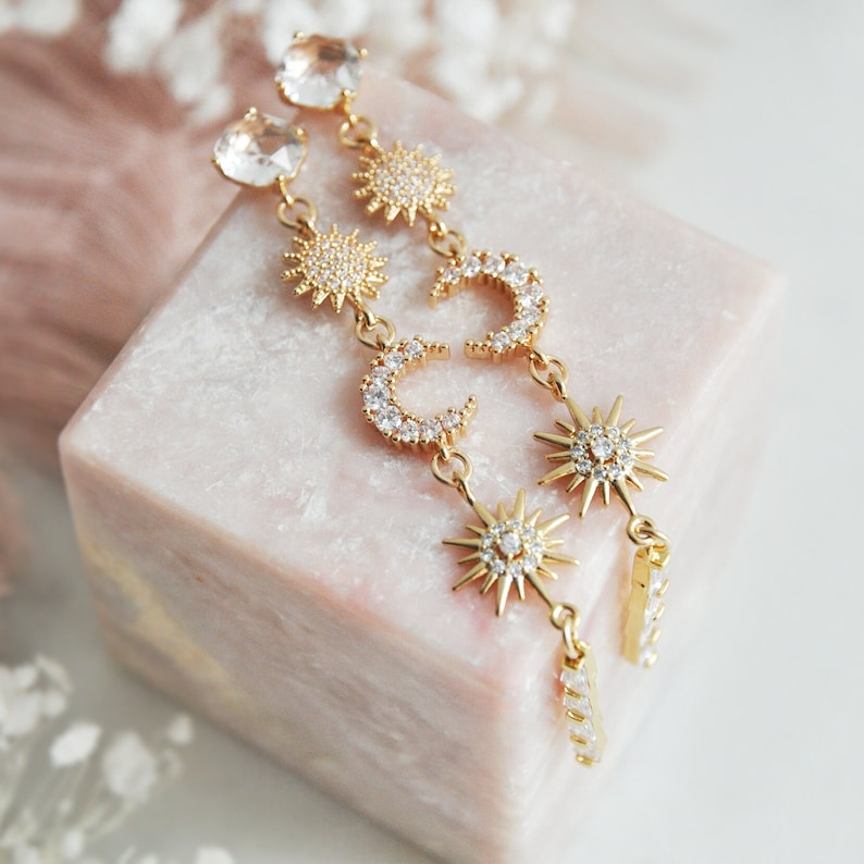 May include: Gold-toned celestial earrings featuring sun and crescent moon designs, adorned with clear crystals. The earrings have a long, dangling style, with a large crystal at the top. The earrings are displayed on a pink, marble-like surface.
