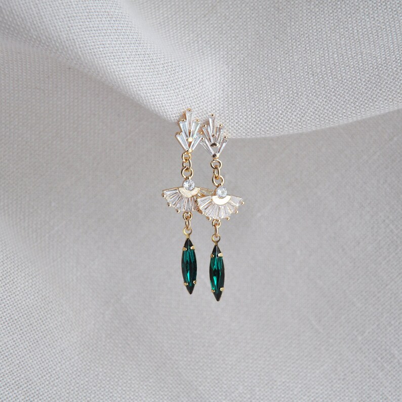 May include: Gold-toned drop earrings with a fan-shaped design, adorned with clear crystals and emerald green teardrop-shaped gems. The earrings have a delicate, elegant appearance, suitable for special occasions.