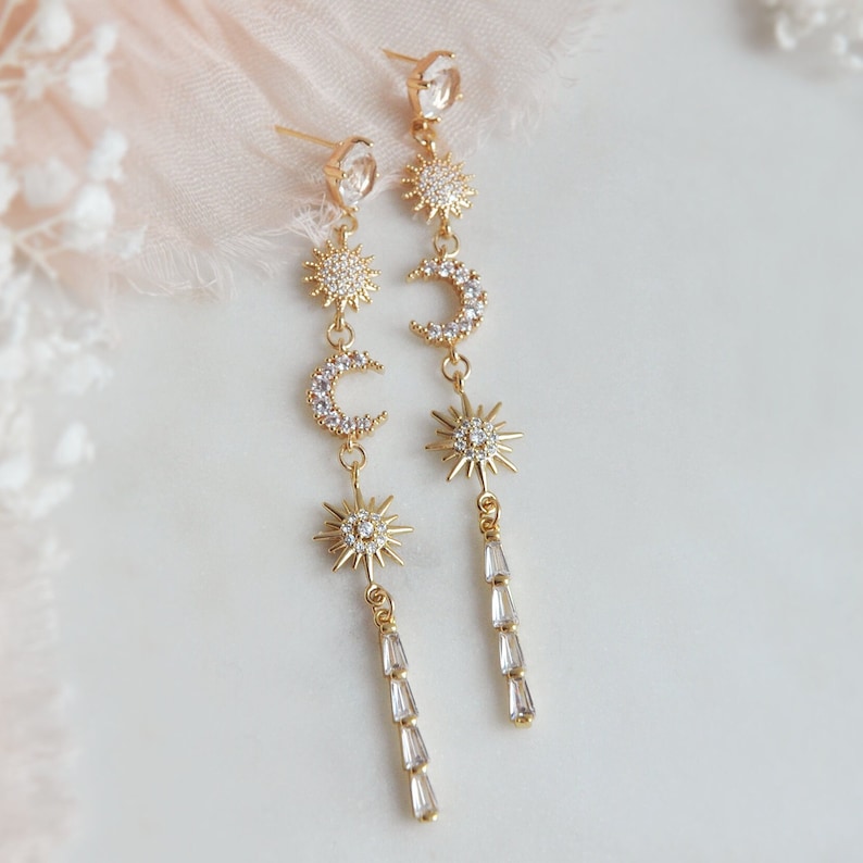 May include: Gold-toned celestial earrings featuring sun and crescent moon charms, adorned with sparkling clear crystals. The earrings have a dangling design with a series of baguette-cut crystals at the bottom. The earrings are designed to be worn with pierced ears.