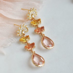 May include: A pair of gold-toned earrings with a sunburst design at the top. The earrings feature yellow, orange, and pink gemstones in a cascading arrangement, culminating in a teardrop-shaped pink gem. The earrings are set against a soft, light background.