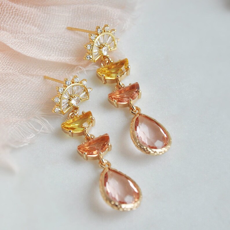 May include: A pair of gold-toned earrings with a sunburst design at the top. The earrings feature yellow, orange, and pink gemstones in a cascading arrangement, culminating in a teardrop-shaped pink gem. The earrings are set against a soft, light background.