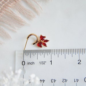 K&ouml;nnte beinhalten: Ein goldfarbener Ohrring mit einem roten Blumenmuster. Der Ohrring hat einen B&uuml;gel und einen Stecker. Die Blume besteht aus roten Edelsteinen. Der Ohrring ist etwa 1,9 cm lang.