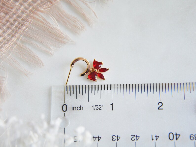 K&ouml;nnte beinhalten: Ein goldfarbener Ohrring mit einem roten Blumenmuster. Der Ohrring hat einen B&uuml;gel und einen Stecker. Die Blume besteht aus roten Edelsteinen. Der Ohrring ist etwa 1,9 cm lang.