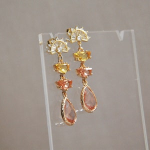 May include: Gold-toned drop earrings with a sunburst design at the top. The earrings feature yellow, orange, and peach-colored gemstones in a tiered arrangement, culminating in a teardrop-shaped peach gem. The earrings are displayed on a clear stand.