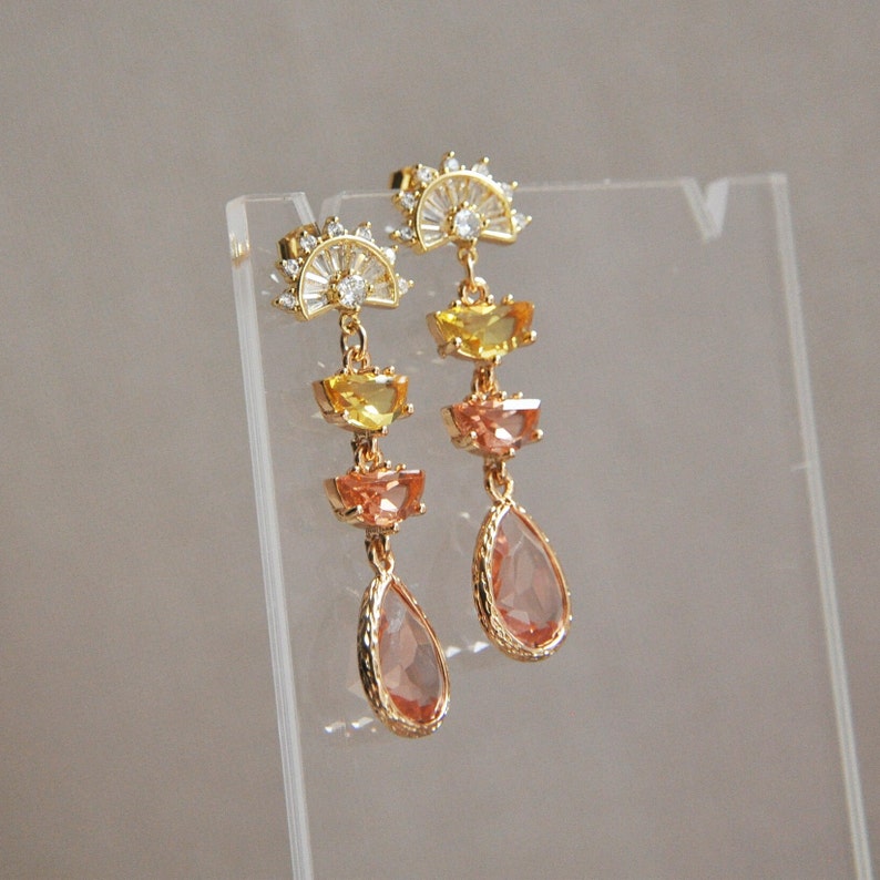 May include: Gold-toned drop earrings with a sunburst design at the top. The earrings feature yellow, orange, and peach-colored gemstones in a tiered arrangement, culminating in a teardrop-shaped peach gem. The earrings are displayed on a clear stand.