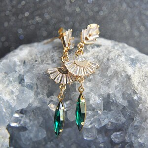 May include: Gold-toned earrings with a fan-shaped design and clear crystals. Each earring features a dangling emerald-green, teardrop-shaped crystal. The earrings are displayed on a light blue, textured surface.