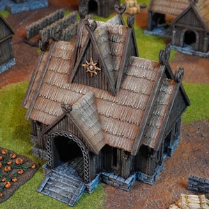 May include: A detailed miniature Viking-style wooden building with a thatched roof. The structure has decorative carvings, a starburst emblem, and a stone foundation. The building is set on a green surface, with other miniature buildings in the background.