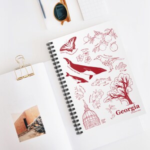 May include: A spiral-bound notebook with a white cover featuring red illustrations of Georgia state symbols, including a butterfly, whale, and peach branch. The text "Georgia the peach state" is also visible. A gold paper clip and sunglasses are in the background.