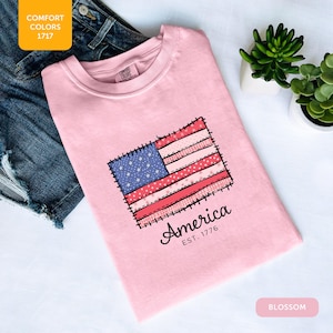 May include: A light pink t-shirt with a stitched American flag design. The flag features blue stars, red and white stripes, and the word "America" with "EST. 1776" below. The shirt is next to denim shorts.