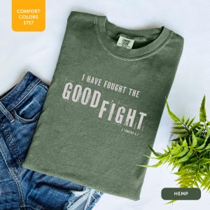 May include: Olive green t-shirt with the text "I HAVE FOUGHT THE GOOD FIGHT" in white, with "2 Timothy 4:7" below. The shirt is folded on a white surface next to blue denim shorts and a fern.