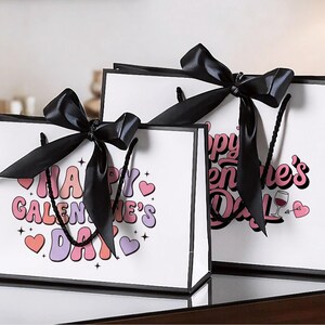 Pu&ograve; includere: Due sacchetti regalo bianchi con bordi e manici neri, ciascuno ornato da un grande fiocco di raso nero. Le borse presentano il testo "Happy Galentine's Day" in un font retr&ograve; colorato, insieme a grafiche a forma di cuore.