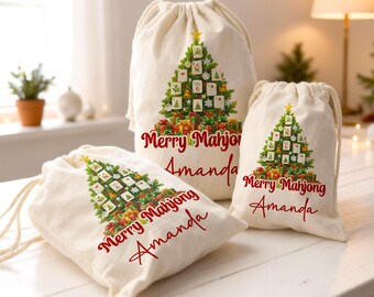 Puede incluir: Tres bolsas con cordón de color crema con un diseño de árbol de Navidad hecho con fichas de Mahjong. Las bolsas tienen el texto "Merry Mahjong Amanda" en rojo. Una estrella dorada corona el árbol, con regalos en la base.