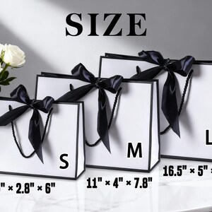 Pu&ograve; includere: Tre sacchetti regalo bianchi con bordi e manici neri, ognuno ornato da un fiocco di raso nero. Le taglie sono S, M e L, con dimensioni di 19cm x 7cm x 15cm, 28cm x 10cm x 20cm e 42cm x 13cm x 30cm. La parola "SIZE" &egrave; in alto.