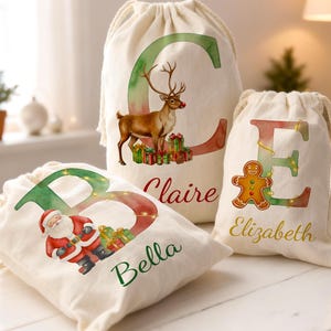 May include: Three off-white drawstring gift bags with Christmas-themed designs. One bag features a reindeer, another Santa Claus, and the third a gingerbread man. Each bag has a letter and a name printed on it.