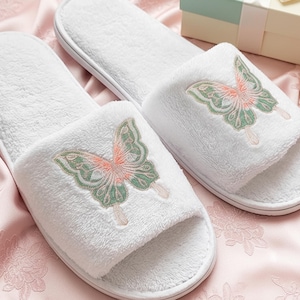 Op de afbeelding: Een paar witte badstof slippers met een geborduurd vlinderontwerp in groen en koraal. De open teenslippers liggen op een roze satijnen oppervlak, wat wijst op een spa- of ontspanningsthema.