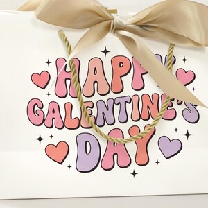 Pu&ograve; includere: Un sacchetto regalo bianco con la scritta "HAPPY GALENTINE'S DAY" in lettere rosa, pesca e viola. La borsa ha un fiocco di nastro dorato e manici in corda. Cuori rosa e viola e stelle nere decorano la borsa.