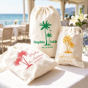 May include: Three off-white drawstring canvas bags featuring palm tree graphics and personalized text. One bag displays green text reading Sophia Isidore 06.12.2028, another shows yellow text reading Sophia & Isidore 06.12.2028, and one shows red text.
