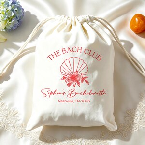 Puede incluir: Una bolsa de tela color crema con cordón y un gráfico rojo de una concha marina con una flor. El texto dice THE BACH CLUB, Sophia's Bachelorette y Nashville, TN-2026 en tinta roja. La bolsa tiene dos cordones de algodón blanco.