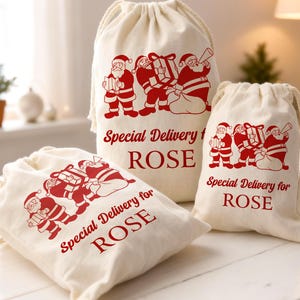 May include: Three off-white drawstring bags with red Santa Claus illustrations and the text "Special Delivery for ROSE". The bags vary in size and are made of a soft, natural fabric, perfect for holiday gifting.