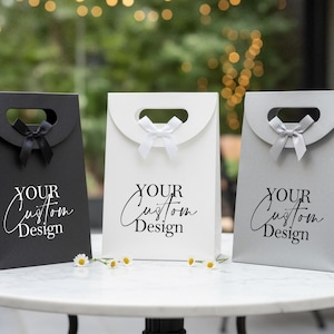 Op de afbeelding: Drie cadeauzakken in zwart, wit en zilver, elk met een handvat en een strik. De tassen hebben de tekst "YOUR Custom Design" op de voorkant gedrukt. Madeliefjes zijn voor de tassen geplaatst.