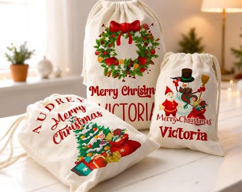 Puede incluir: Tres bolsas de regalo navideñas de color crema con cordón. Una bolsa presenta una corona con un lazo rojo, el texto "Merry Christmas" y el nombre "Victoria". Otra bolsa tiene un árbol de Navidad y regalos, con el nombre "Audrey". La tercera bolsa tiene un muñeco de nieve y Papá Noel con el nombre "Victoria".