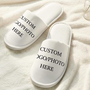 Op de afbeelding: Twee witte slippers met open teen met de tekst "CUSTOM LOGO/PHOTO HERE" in zwart gedrukt. De slippers liggen op een wit, getextureerd oppervlak, wat wijst op een spa- of hotelomgeving. Het ontwerp maakt personalisatie mogelijk.