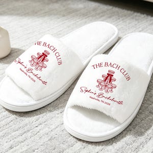 Puede incluir: Un par de pantuflas blancas de felpa con punta abierta que presentan un gráfico granate de una torre de copas de champán con un lazo. El texto dice THE BACH CLUB, Sophia's Bachelorette y Nashville, TN-2026 sobre la tela suave.