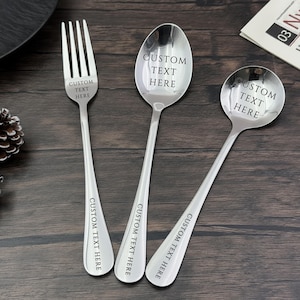 May include: A stainless steel fork, spoon, and ladle set with the text "CUSTOM TEXT HERE" engraved on each utensil. The utensils are on a dark wooden surface, next to a black plate and a pinecone.