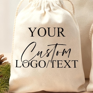 以下が含まれることがあります： 黒い文字で「YOUR Custom LOGO/TEXT」と書かれた、クリーム色の巾着袋。袋はテクスチャード加工された生地で作られています。松ぼっくりと緑が背景にあります。