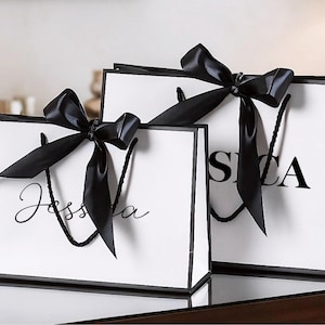 Può includere: Due sacchetti regalo bianchi con bordi e manici neri. Ogni sacchetto ha un grande fiocco di raso nero. Un sacchetto ha il nome "Jessica" in corsivo, l'altro "SICA" in grassetto.