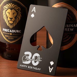 Puede incluir: Un abrebotellas de metal negro con forma de carta de juego, con el As de Picas y un recorte en forma de corazón. La tarjeta está personalizada con el nombre "Charlotte", el número "30" y las palabras "Happy Birthday". Dos botellas de cerveza en el fondo.