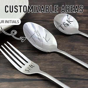 Könnte beinhalten: Edelstahl-Besteck-Set mit eingravierten Initialen. Das Set enthält eine Gabel und zwei Löffel, jeweils mit einer anderen Initialenkombination. Der Text "CUSTOMIZABLE AREAS" steht oben, mit "YOUR INITIALS" in einem weißen Oval. Das Besteck liegt auf einer dunklen Holzoberfläche.