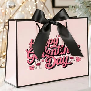 Pu&ograve; includere: Una borsa regalo rosa con bordi neri e un grande fiocco nero. La borsa presenta la scritta "Happy Valentine's Day" in rosa e bianco, insieme a illustrazioni di cuori e calici di vino. La borsa &egrave; appoggiata su una superficie bianca.