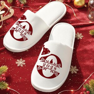 May include: A pair of white, open-toe slippers with a red design featuring the year "2028" and a Christmas tree. The slippers are on a red surface with holiday decorations, including greenery and lights.