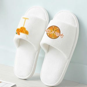 May include: A pair of white spa slippers with orange and yellow cocktail graphics. One slipper features a cocktail illustration, and the other has a graphic of an orange slice with the text "Spritz-O-Glo".