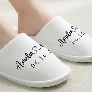 以下が含まれることがあります： 白くふわふわのスリッパ。各スリッパには「Amelia」という名前と「06.16」という日付が黒い筆記体で印刷されています。スリッパはシンプルなスリップオンデザインで、白いソールが付いています。画像には2つのスリッパが写っています。
