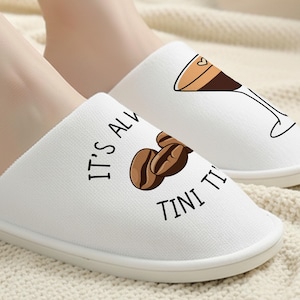 Peut inclure: Des chaussons blancs à enfiler avec des motifs sur le thème du café. Un chausson présente des grains de café et le texte "IT'S ALWAYS TINI TI". L'autre chausson a une illustration de cocktail au café. Les chaussons sont sur une surface crème.