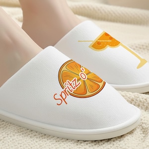May include: White slip-on slippers with orange and yellow cocktail-themed graphics. One slipper features a slice of orange with the word "Spritz" and the other has a cocktail glass with an orange slice.