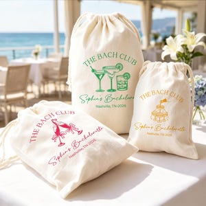 May include: Three off-white cotton drawstring bags with printed graphics. The largest features green cocktail illustrations and text reading THE BACH CLUB, Sophia's Bachelorette, Nashville, TN-2026. Smaller bags feature pink and gold designs.