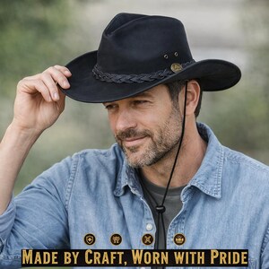May include: A black cowboy hat with a braided band and a chin strap. The hat has a wide brim and is worn by a person wearing a denim shirt. The text "MADE BY CRAFT, WORN WITH PRIDE" is at the bottom of the image.