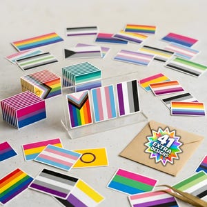 Peut inclure: Une collection d'autocollants rectangulaires représentant des drapeaux de fierté aux motifs variés, incluant des piles et des pièces éparses. Un support en acrylique transparent présente trois autocollants. Une enveloppe brune porte le texte 41 EXTRA DESIGNS.