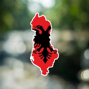Puede incluir: Una pegatina con la forma del mapa de Albania, que presenta un fondo rojo vibrante con una silueta negra de un águila bicéfala centrada dentro de los bordes. El diseño está delineado con un borde blanco fino, creando un contraste.