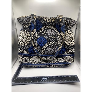 Puede incluir: Un bolso tote con un estampado floral negro, azul y blanco. El bolso tiene un bolsillo frontal y dos asas. El bolso está hecho de un material acolchado. El bolso mide aproximadamente 38 cm de ancho.