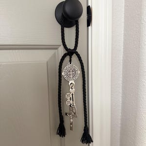 Puede incluir: Colgador de puerta de cuerda negra con un colgante decorativo plateado con un emblema religioso, una llave y un crucifijo. El colgador está sujeto a un pomo de puerta negro en una puerta gris.