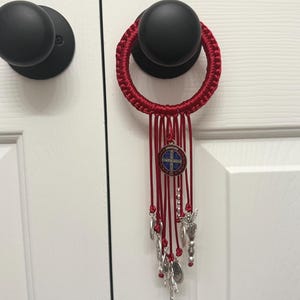 May include: A red macrame door hanger with dangling red cords, silver charms, and a medallion. The medallion has a blue and white design. The door hanger is hanging on a black doorknob.